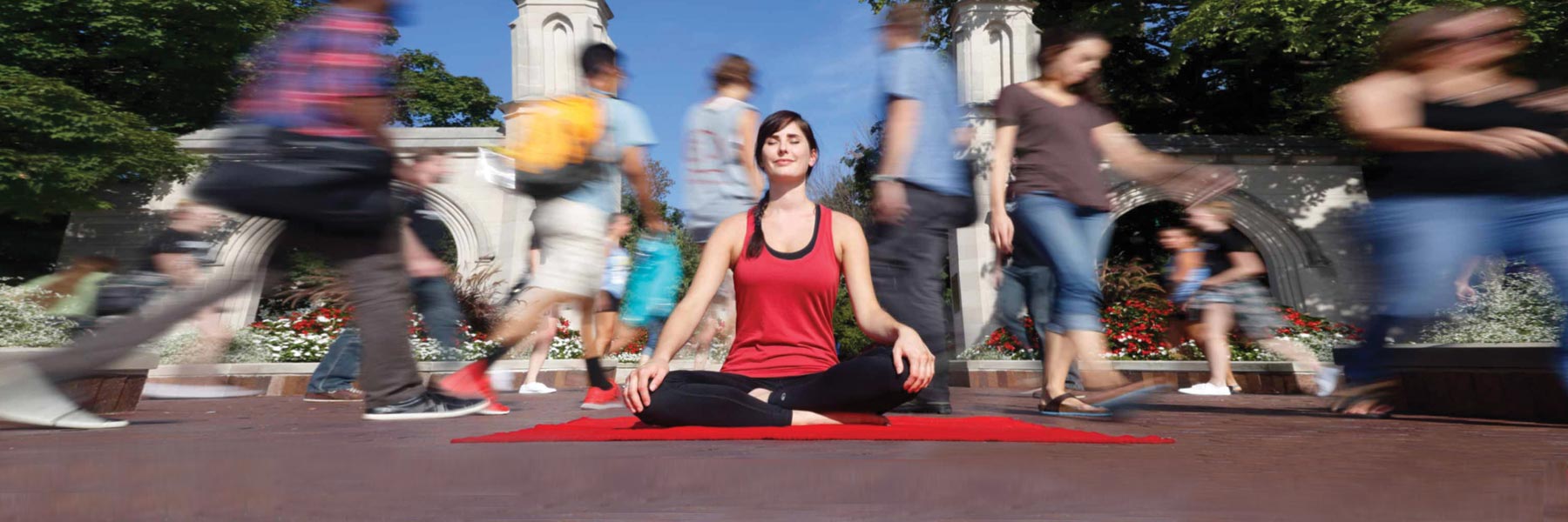 A woman sitting in lotus on a yoga mat in front of the sample gates while people rush past her