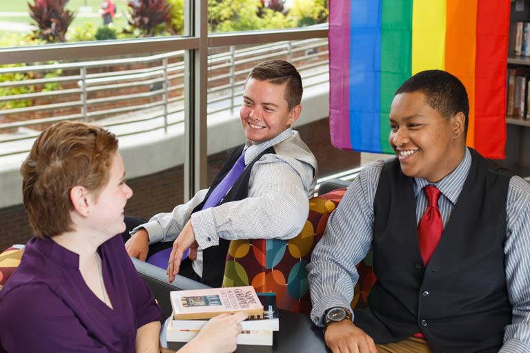 three people smiling at each other in front of a pride rainbow flag